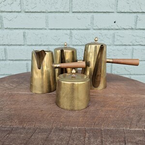 May include: A vintage brass coffee set, including a creamer, sugar bowl, and two coffee pots. The set features wooden handles and lids with small finials. The pieces are arranged on a wooden surface against a light blue brick wall.