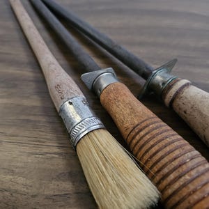 May include: Close-up of three antique kitchen tools: a brush with a wooden handle and metal ferrule, and two steel sharpening rods with wooden handles. The tools are arranged on a dark wooden surface.
