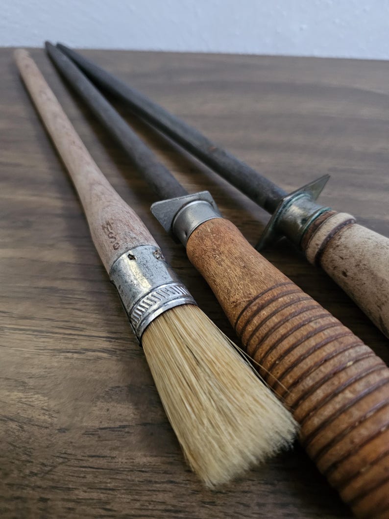 May include: Close-up of three antique kitchen tools: a brush with a wooden handle and metal ferrule, and two steel sharpening rods with wooden handles. The tools are arranged on a dark wooden surface.