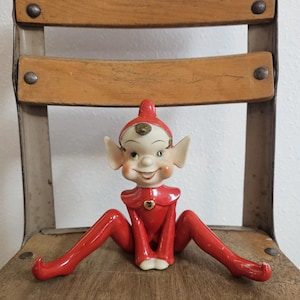 May include: A vintage ceramic elf figurine with a red outfit and a smiling face. The elf is sitting on a wooden chair with a metal frame. The elf has a red hat and a gold heart on its chest.