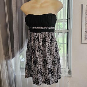 May include: A strapless dress on a mannequin. The top is black, with a ruched design. The waist is accented with a black band. The skirt features a black and white floral lace pattern. The dress is knee-length.