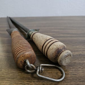 May include: Two vintage knife sharpeners with wooden handles. One handle is ribbed, the other is smooth with dark bands. The metal rods are silver and extend from the handles. A metal loop connects to the ribbed handle.