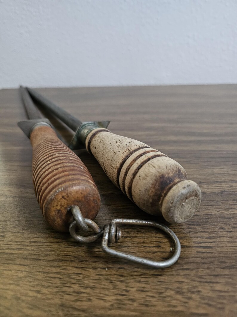 May include: Two vintage knife sharpeners with wooden handles. One handle is ribbed, the other is smooth with dark bands. The metal rods are silver and extend from the handles. A metal loop connects to the ribbed handle.