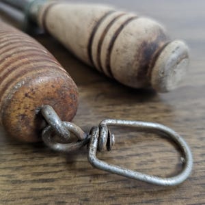 May include: Close-up of two vintage metal and wood tools. One tool has a wooden handle with dark brown rings, and a metal loop. The other tool has a light brown wooden handle. The tools are on a wooden surface.