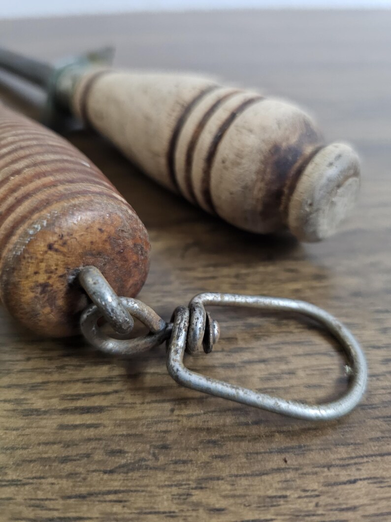May include: Close-up of two vintage metal and wood tools. One tool has a wooden handle with dark brown rings, and a metal loop. The other tool has a light brown wooden handle. The tools are on a wooden surface.