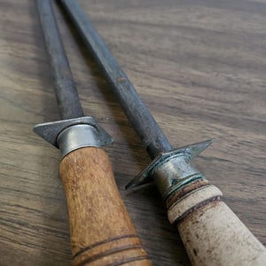 May include: Two steel knife sharpening rods with wooden handles. One handle is light brown with a ribbed design, the other is a lighter color with a smooth finish. The rods are angled and rest on a wooden surface.