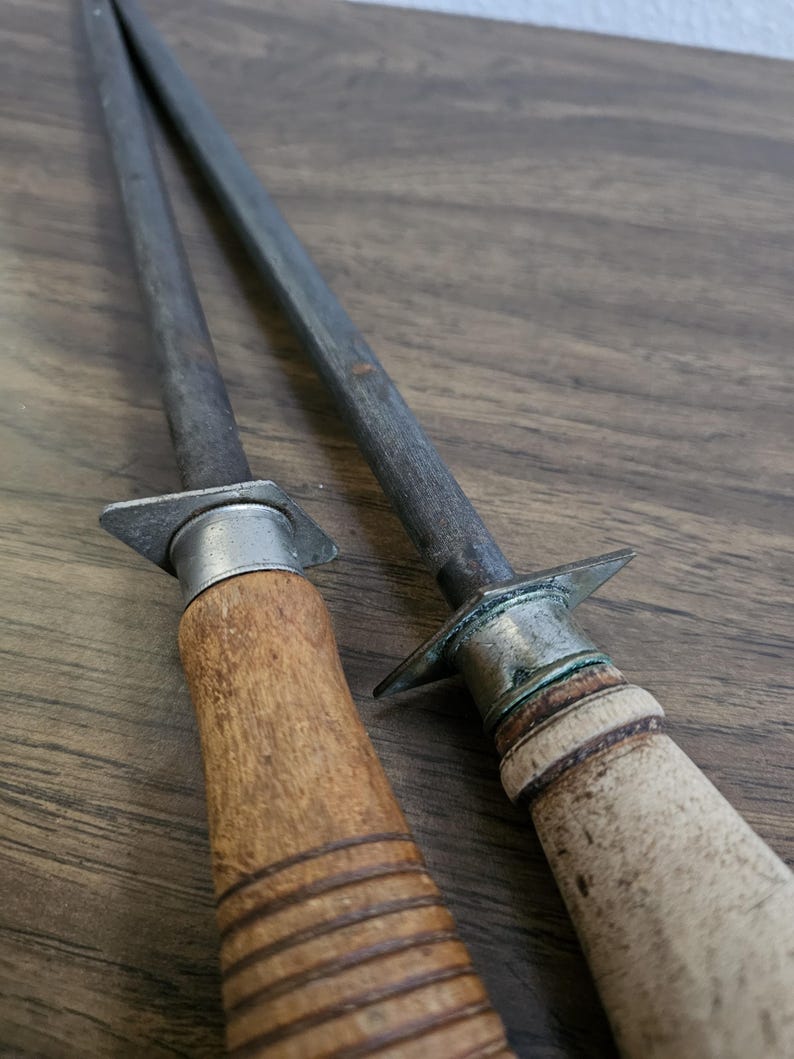 May include: Two steel knife sharpening rods with wooden handles. One handle is light brown with a ribbed design, the other is a lighter color with a smooth finish. The rods are angled and rest on a wooden surface.