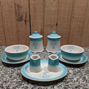 May include: A set of blue and white ceramic teacups, saucers, and creamer pitchers with Chinese characters. The set includes two sugar bowls with lids, two creamer pitchers, two teacups, and two saucers.