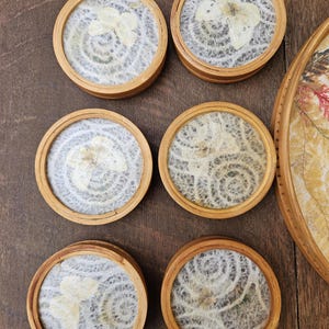 K&ouml;nnte beinhalten: Set aus sechs runden Untersetzern mit einem Rahmen aus Naturholz. Jeder Untersetzer zeigt ein Design mit getrockneten Blumen auf einem wei&szlig;en, strukturierten Hintergrund. Die Untersetzer sind auf einer Holzoberfl&auml;che angeordnet und zeigen ihre dekorative und funktionale Natur.