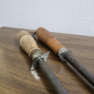 May include: Two vintage steel knife sharpeners with wooden handles. One handle is light-colored with a ridged design, the other is darker with a grooved pattern. The metal rods are dark gray, and the tools rest on a wooden surface.