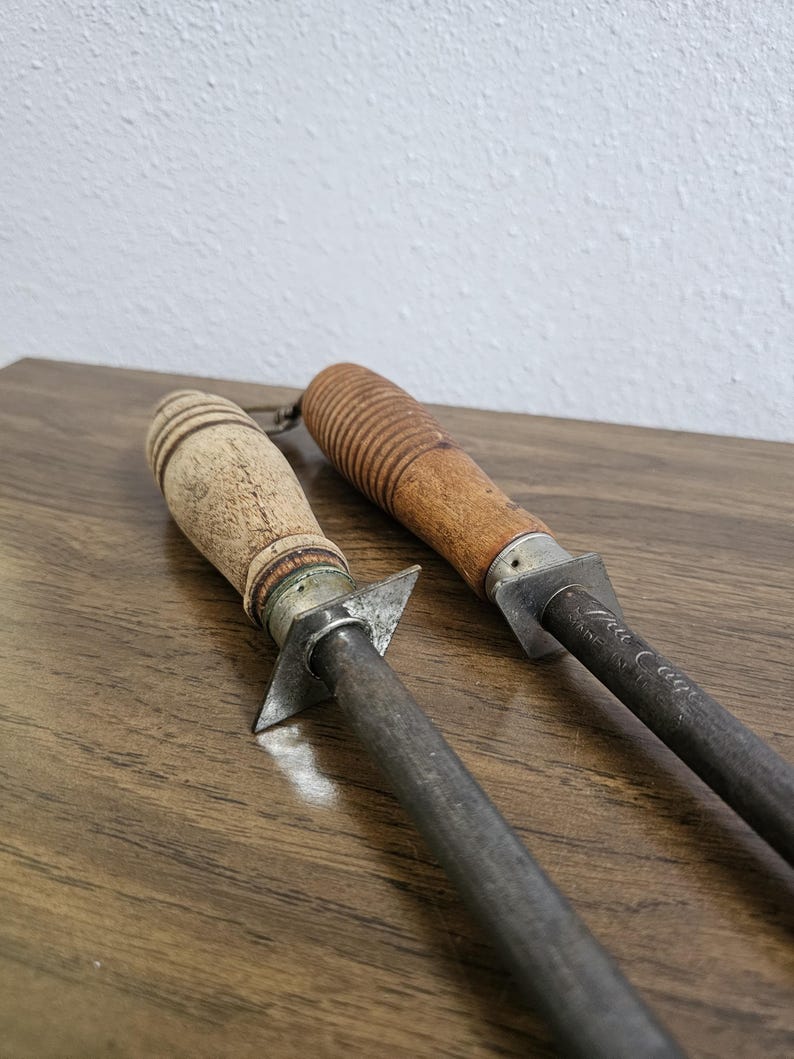 May include: Two vintage steel knife sharpeners with wooden handles. One handle is light-colored with a ridged design, the other is darker with a grooved pattern. The metal rods are dark gray, and the tools rest on a wooden surface.