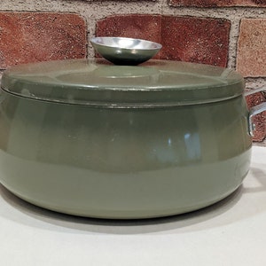May include: An olive green cooking pot with a lid and two silver handles. The lid has a small, silver, bowl-shaped handle. The pot is round and sits on a white surface. The background is a brick wall.