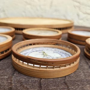 K&ouml;nnte beinhalten: Eine Sammlung runder Bambus-Untersetzer mit einem hellen, von der Natur inspirierten Design in der Mitte. Die Untersetzer haben eine nat&uuml;rliche Holzoberfl&auml;che und einen dekorativen, geschlitzten Rand. Ein gro&szlig;er, runder Bambusteller befindet sich im Hintergrund.