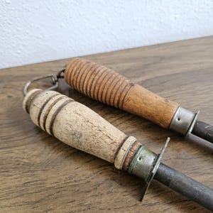 May include: Two vintage knife sharpeners with wooden handles. One handle is light-colored, the other is a darker wood. Both have metal bolsters and steel sharpening rods. The handles show signs of wear.