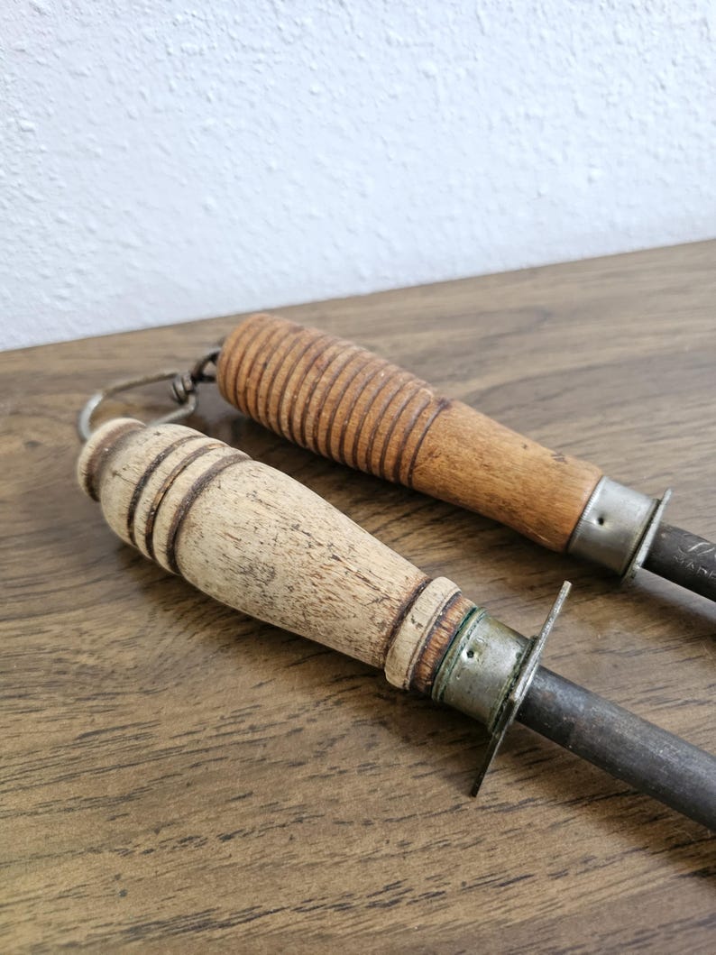 May include: Two vintage knife sharpeners with wooden handles. One handle is light-colored, the other is a darker wood. Both have metal bolsters and steel sharpening rods. The handles show signs of wear.