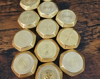 Könnte beinhalten: Eine Sammlung von zehn goldfarbenen Metallknöpfen, die jeweils ein kreisförmiges Design mit einem zentralen Buchstaben R, dem Wort CANADA und der Jahreszahl 1939 auf der Oberfläche aufweisen. Die Knöpfe haben einen facettierten Rand und ein Metallfinish.