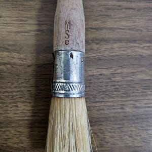 May include: Close-up of a paint brush with a wooden handle and natural bristles. The handle has the letters "WSE" stamped on it. The brush is held together with a silver metal ferrule.