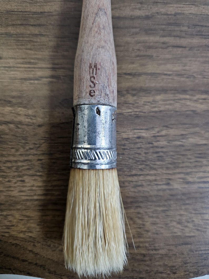 May include: Close-up of a paint brush with a wooden handle and natural bristles. The handle has the letters "WSE" stamped on it. The brush is held together with a silver metal ferrule.