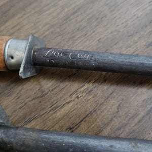May include: Close-up of a steel knife sharpener with a wooden handle. The text "Tru Edge" and "Made in U.S.A." are visible on the metal. The sharpener is resting on a wooden surface.