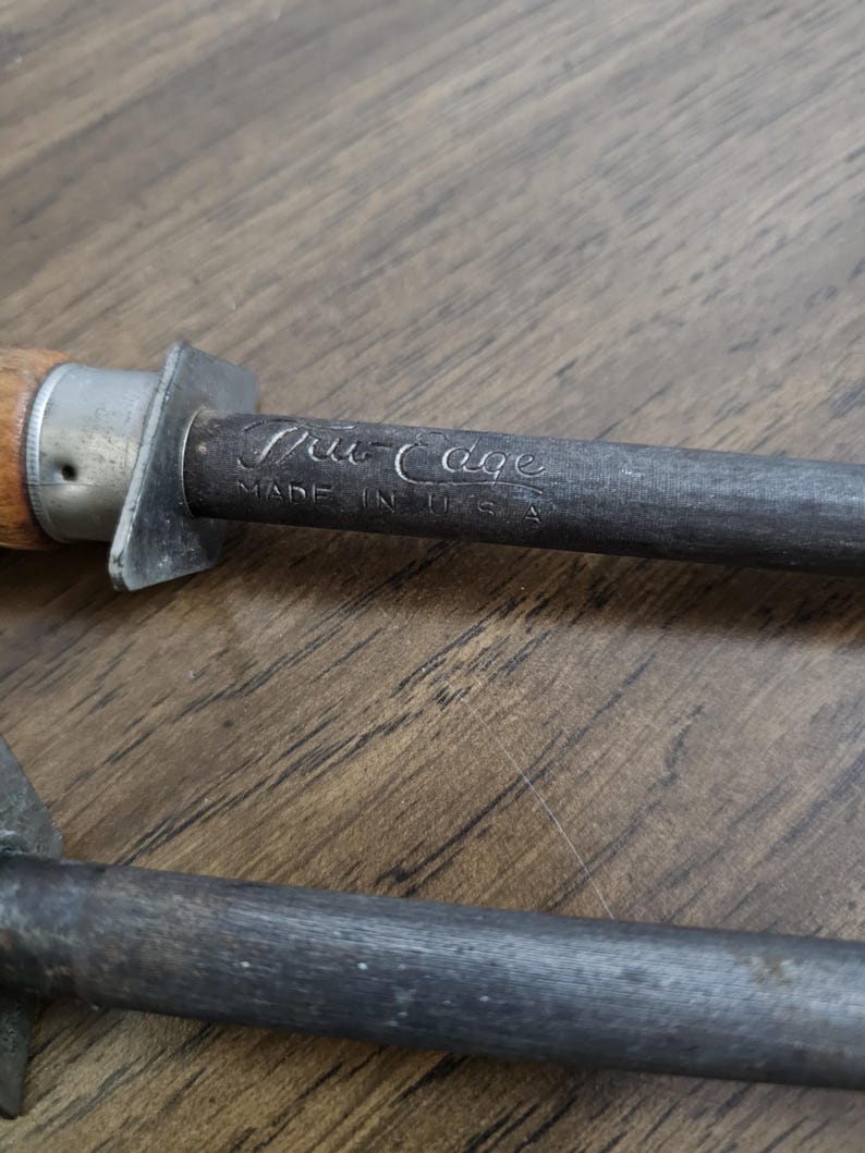 May include: Close-up of a steel knife sharpener with a wooden handle. The text "Tru Edge" and "Made in U.S.A." are visible on the metal. The sharpener is resting on a wooden surface.
