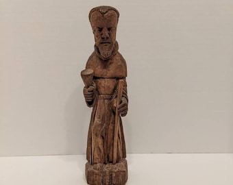 Könnte beinhalten: Eine handgeschnitzte Holzfigur eines bärtigen Mannes in einer Kapuzenrobe. Die Figur hält einen kleinen Kelch in der rechten Hand und einen langen Wanderstab in der linken Hand. Die Skulptur hat eine dunkelbraune Oberfläche und steht auf einem Sockel.