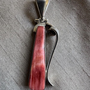 May include: A vertical pendant featuring a polished silver frame and a long, tapered inlay of mottled pink and white stone. The silver setting includes a large, triangular bail at the top and a curved, decorative silver wire accent on the right side.
