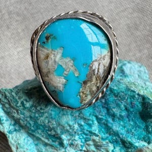 Könnte beinhalten: Ein Silberring mit einem großen, ovalen Türkisstein. Der hellblaue Stein hat weiße und braune Einschlüsse. Die Fassung des Rings hat ein gedrehtes Silberseildesign.