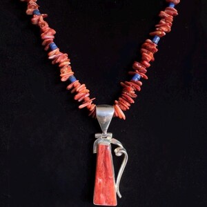 May include: A necklace featuring a silver pendant with a red rectangular stone and a curved silver accent. The necklace has a strand of red coral chips interspersed with blue beads, set against a black background.