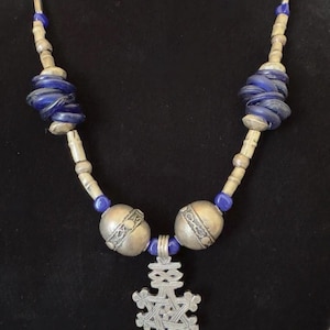 May include: A necklace featuring a silver-toned metal cross pendant with intricate geometric engravings. The chain consists of alternating silver-toned cylindrical beads, small blue glass beads, and two larger clusters of stacked blue glass discs.