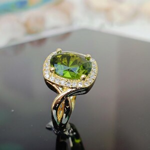 May include: A gold ring featuring a large, oval-cut, olive-green gemstone surrounded by small, clear gemstones. The ring's band has a unique, swirling design. The ring is displayed on a reflective black surface.