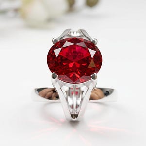 May include: A silver ring with a large, oval-cut, deep red gemstone. The gemstone is the focal point, held by four prongs. The ring band is smooth and polished, reflecting light. The setting is modern and elegant.