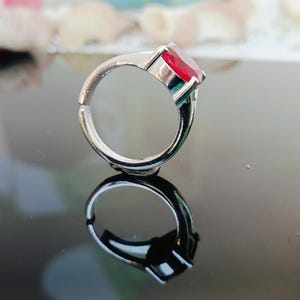 May include: A silver-toned ring with a large, square-cut, red gemstone. The ring is set on a reflective surface, creating a mirrored image. The ring's band is adjustable. The gemstone is the focal point of this jewelry piece.