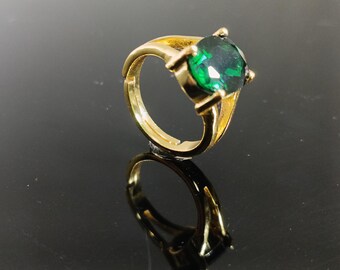 May include: A gold-toned ring featuring a large, emerald-green gemstone. The ring's band has a classic design, and the gemstone is securely held in place by four prongs. The ring is displayed on a reflective surface.