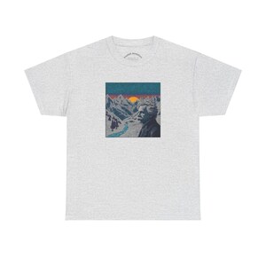Könnte beinhalten: Hellgraues T-Shirt mit einem quadratischen Aufdruck eines Mannes vor einer Berglandschaft mit Fluss und Sonnenuntergang. Die Grafik ist in Blau-, Orange- und Grautönen gehalten. Das Shirt hat kurze Ärmel und einen Rundhalsausschnitt.