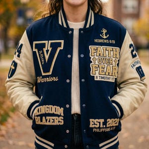 May include: Navy blue and tan varsity jacket with the text "V Warriors" and "Faith Over Fear". The jacket has a button-up front, ribbed cuffs, and a striped collar. The sleeves are tan, and the body is navy blue. The jacket also has the text "Kingdom Blazers" and "Est. 2021".