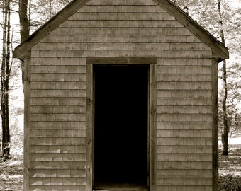May include: A small, wooden shed with a single door. The shed is made of wood shingles and has a simple, rustic design. The door is open, revealing a dark interior.