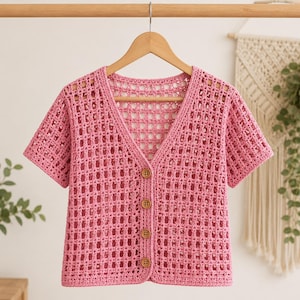 May include: A short-sleeved pink crochet cardigan featuring an open grid pattern, a V-neckline, and four round brown buttons down the centre front. The garment is displayed on a wooden hanger against a neutral background.