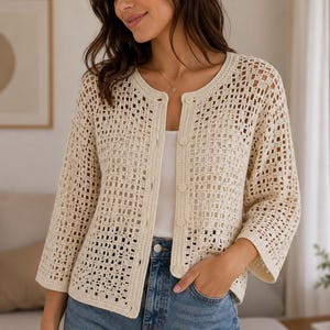 Può includere: Un cardigan all'uncinetto color crema a maglia aperta, caratterizzato da scollo rotondo, maniche a tre quarti e chiusura frontale con bottoni. Il capo presenta un motivo a griglia quadrata e un bordo a coste solido lungo l'apertura.