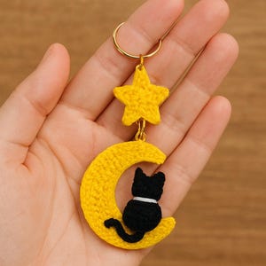 Op de afbeelding: Handgemaakte gele gehaakte sleutelhanger met een halve maan met een zwarte kat en een gele ster. De sleutelhanger heeft een goudkleurige ring om aan sleutels of tassen te bevestigen. De kat heeft een witte halsband.