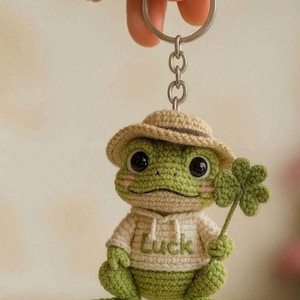 Pode incluir: Chaveiro de sapo de crochê em tons de verde e bege. O sapo usa um moletom bege com a palavra "Luck" e um chapéu a condizer. Ele segura um trevo de quatro folhas. O chaveiro é preso a um anel de metal prateado.