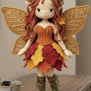 May include: A handmade crochet fairy doll in autumnal hues. The doll has a cream face, red-orange hair, and a dress of crochet leaves in red, orange, and yellow. It has large, leaf-shaped wings and brown boots with green laces. The doll is approximately 25 cm tall.