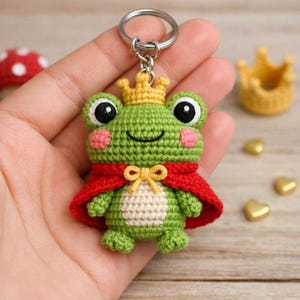 Pode incluir: Um chaveiro de sapo verde de crochê com uma coroa dourada e uma capa vermelha. O sapo tem olhos grandes pretos, bochechas rosadas e uma boca sorridente. O chaveiro está preso a um anel de metal prateado.