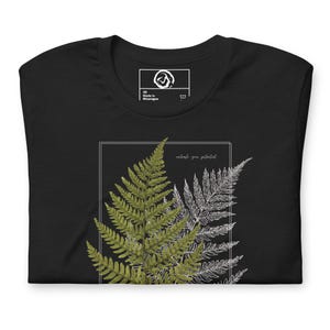 Könnte beinhalten: Schwarzes T-Shirt mit einem Farn-Design in Grün und Weiß, umrahmt von einem Rechteck. Der Text "unleash your potential" ist in zarter Schrift geschrieben. Das Shirt hat ein Etikett mit der Aufschrift "XS Made in Nicaragua".