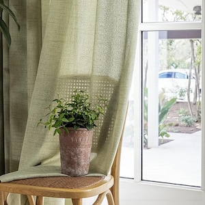 May include: Pale green linen curtains draped over a wooden chair with a woven seat. A terracotta pot with a green plant sits on the chair. A window with a view of a white car and greenery is in the background.