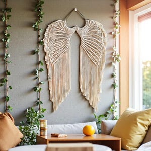 Puede incluir: Un gran colgante de pared con alas de ángel de macramé beige. Las alas están hechas de cordón de algodón de color natural tejido y anudado, con un diseño detallado de plumas. La decoración de la pared está suspendida de un lazo de metal y cuelga de una pared gris.