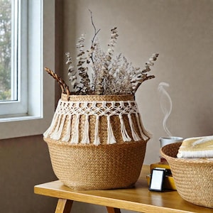 Könnte beinhalten: Ein geflochtener Korb mit Makramee-Besatz und Quasten, gefüllt mit getrockneten Pflanzen. Der Korb hat zwei Griffe und steht auf einem Holztisch. Ein kleinerer Korb ist im Hintergrund zu sehen.
