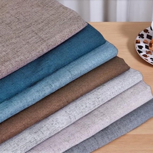 May include: A selection of fabric swatches in shades of beige, teal, brown, and grey, with a textured finish. A teacup and saucer with a playing card design sit on a wooden surface.