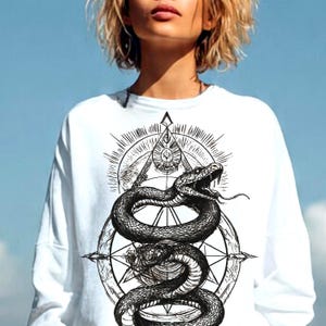 Op de afbeelding: Een wit shirt met lange mouwen en een zwarte grafische print op de borst. Het ontwerp toont een kronkelende slang met een open bek en uitgestoken tong, geplaatst over een geometrische kompasachtige ster en een driehoekige zon.