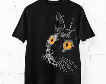 Può includere: Una t-shirt nera a maniche corte con un'illustrazione a linee bianche di una testa di gatto di profilo. Il gatto ha grandi occhi arancioni brillanti e baffi dettagliati, con il disegno centrato sulla parte anteriore del capo.