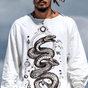 Puede incluir: Una camiseta blanca de manga larga con una detallada ilustración en tinta negra de una serpiente enroscada con la boca abierta. El gráfico incluye formas geométricas, motivos circulares y pequeños detalles en forma de estrella alrededor.